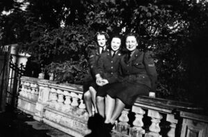 Black and white image circa 1944-46 of three women in military uniform sitting on balustrade in front of group of trees.