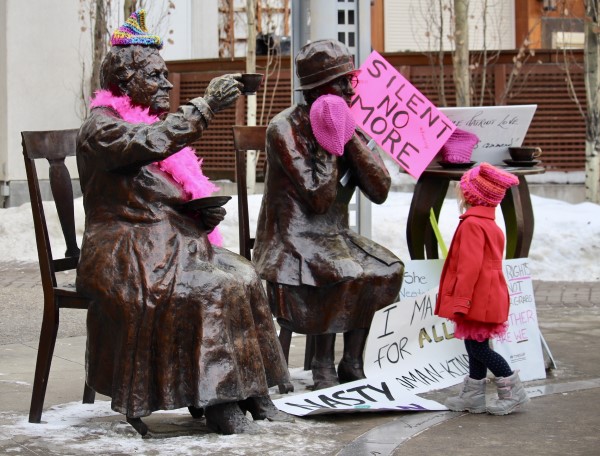 Two bronze statues of women sitting down with hands raising teacups. They are wearing pink knitted wool hat and are surrounded by Women's March posters. A small child in red coat and pink hat stands in front of them.