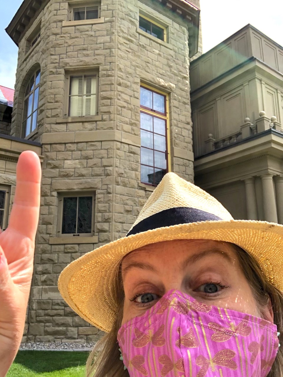Woman wearing a yellow hat and pink face mask pointing at a sandstone building.