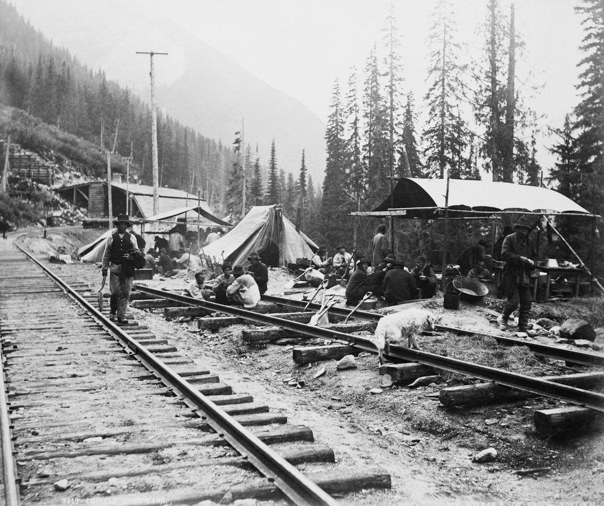 Black and white photo circa 1889 of Chinese work gang on railroad. There is a set of railroad tracks going from bottom right to top left, with tents on far side of tracks. About 30 men are gathered around the tents.